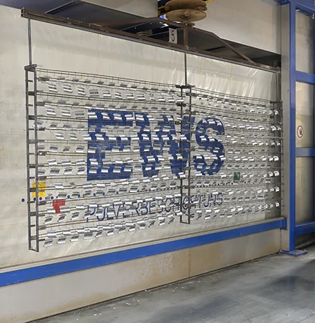 Warengestell mit Aluminiumteilen, die das EWS-Logo bilden, in der Beschichtungsanlage in Vlotho-Exter